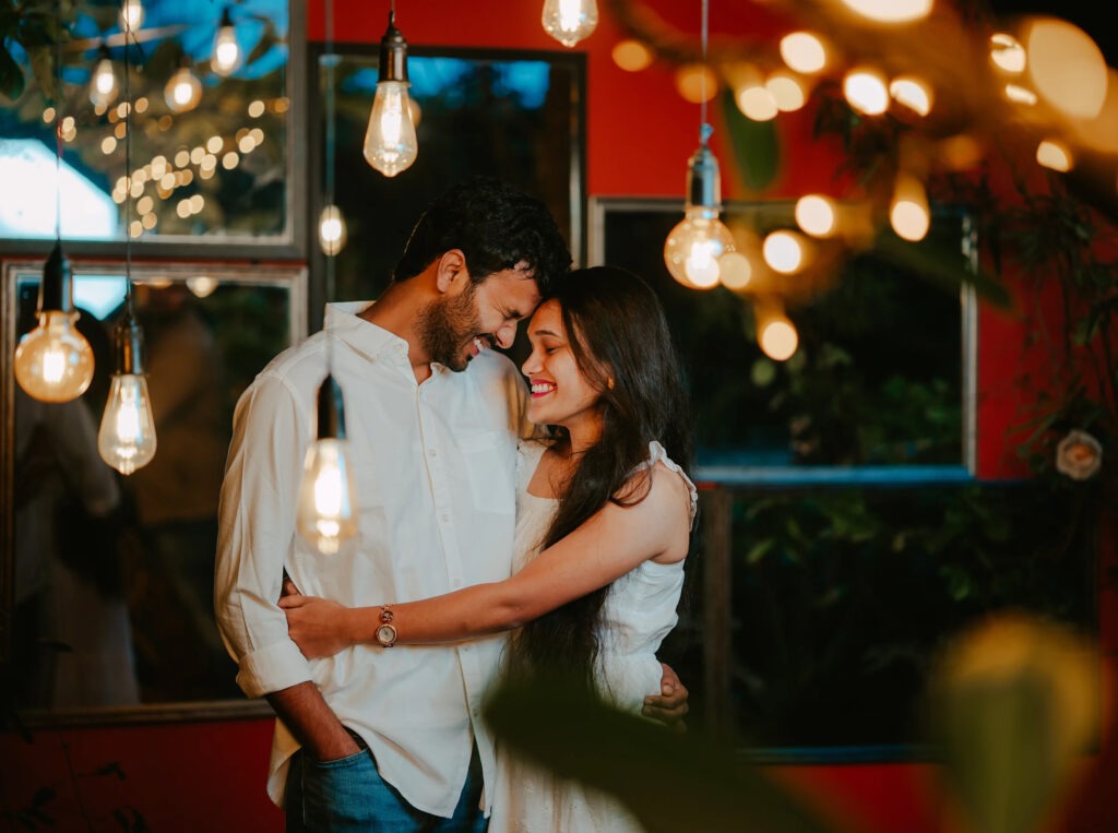 Current image: Engaged couple smiling and embracing during a romantic engagement photoshoot in Guntur with warm lighting and decorative hanging bulbs.