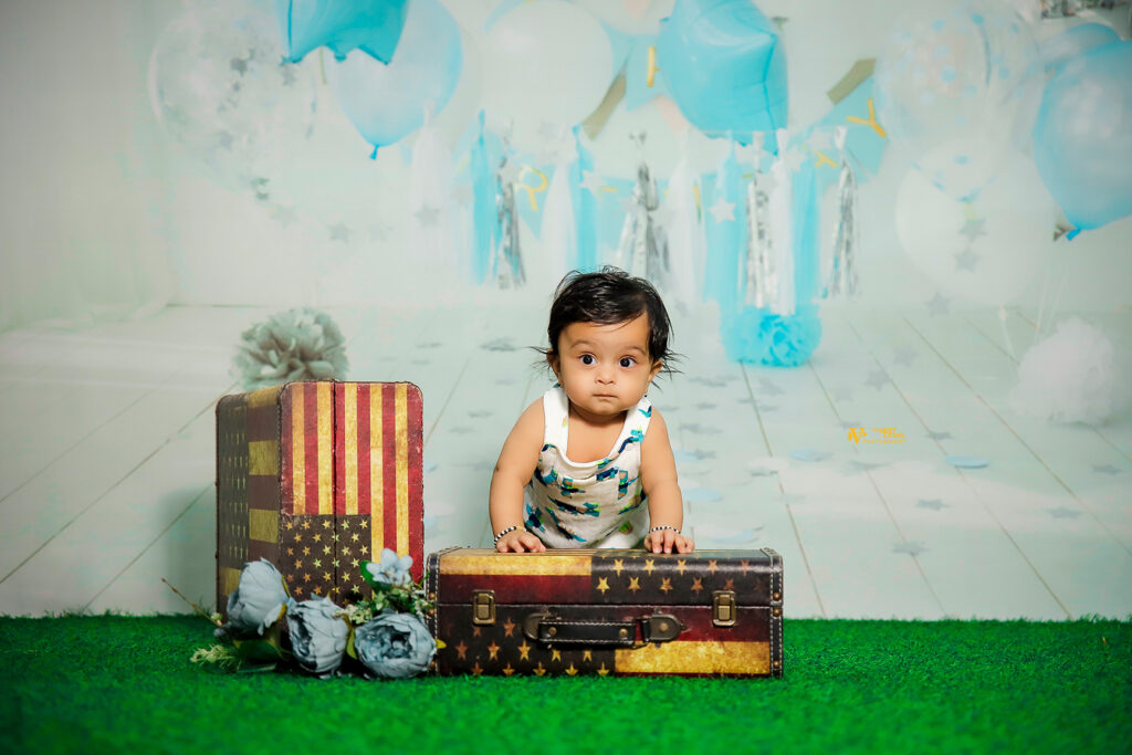 Adorable baby boy standing with support on a vintage suitcase in a blue-themed birthday photoshoot setup with balloons and star decorations – best baby photography in Guntur by Wedlock Jnr.