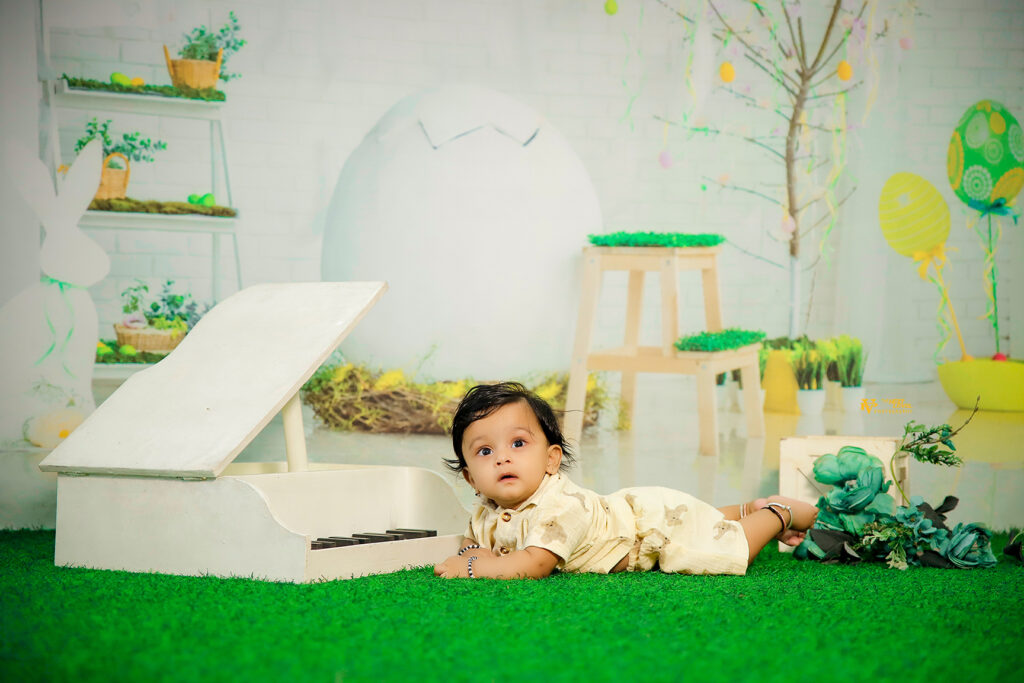 Baby lying on artificial grass near a white toy piano in a spring-themed studio with Easter eggs, cracked egg prop, and colorful decorations – baby photoshoot by best photographers in Guntur at Wedlock Jnr.